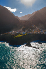 Aerial view of a cliff hugging village on Madeira Island, Portugal, near Seixal or Porto Moniz, with terraced fields, basalt headlands, sea stacks, and dappled light. © Aerial Film Studio