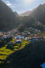 High aerial view shows a whitewashed village with terracotta roofs and terraces on Madeira Island, Portugal, late afternoon light casts shadows toward the Atlantic. © Aerial Film Studio
