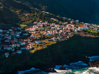 White houses with red roofs line steep terraces above dark cliffs on Madeira, Portugal. Warm late afternoon light reveals rugged slopes and an ocean surf along the north coast. © Aerial Film Studio