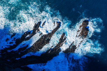 Aerial view of jagged sea stacks in Madeira, Portugal as waves crash. Dark ridges cast long shadows in bright daylight, revealing basalt striations and deep cobalt channels.