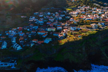 Aerial view of a cliffside village on Madeira Island, Portugal, white houses with terracotta roofs on terraced slopes above the Atlantic, low sun and long shadows. © Aerial Film Studio