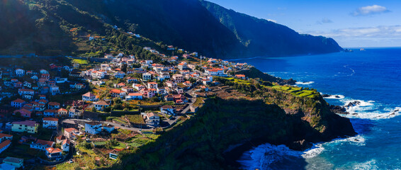 Aerial view shows a cliffside village on Madeira Island, Portugal, white houses and terracotta roofs on green terraces by dark cliffs and deep blue Atlantic at midday.