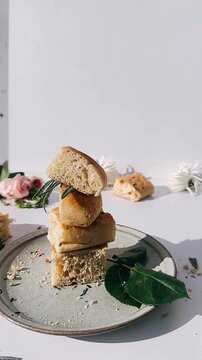 rustic herb focaccia bread pieces stacked on a ceramic plate with scattered crumbs, green leaves, and soft floral accents in natural light.