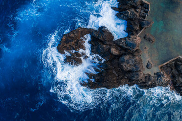 Obraz premium Aerial waves hit jagged volcanic rocks by a stone edged sea pool at Madeira, Portugal. Turquoise to deep blue water churns in bright daylight with high contrast.