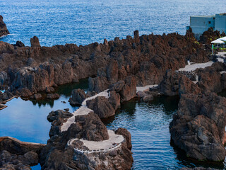 Jagged lava rocks encircle emerald tidal pools and white paths at Porto Moniz, Madeira, Portugal. Late day light and deep blue tones highlight textures and calm water. © Aerial Film Studio