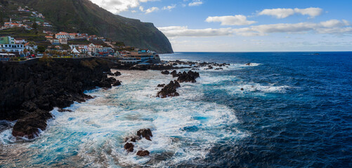 Daylight panoramic view of Porto Moniz, Madeira, with waves striking black lava rocks, whitewashed houses on a slope, a towering headland, and Atlantic waters under clouds. © Aerial Film Studio