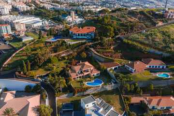 Aerial view of Funchal, Madeira, Portugal shows hillside villas, curving roads, teardrop and kidney pools, and a large estate with blue pool in warm light.