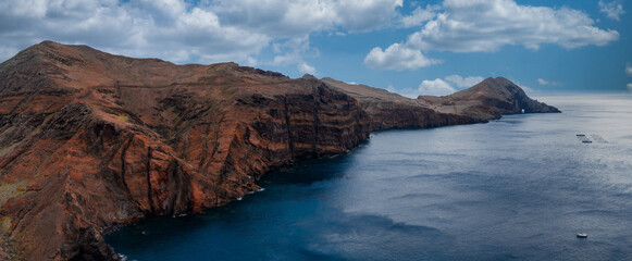Aerial wide view shows stratified reddish cliffs and basalt layers at Ponta de Sao Lourenco, Madeira, Portugal, with calm deep blue water and small boats by day.