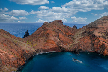 Aerial view shows rust red cliffs at Ponta de Sao Lourenco, Madeira, Portugal, a tour boat crossing turquoise water, sea stacks on the horizon, and bright midday light.