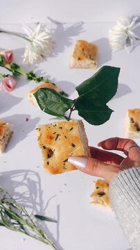 A small square of golden herb focaccia is gently held by a hand, accented with a fresh green leaf against a clean white surface and soft shadows.