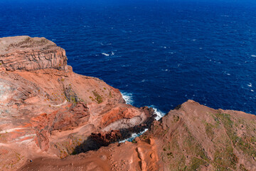 Aerial view shows rust red cliffs at Ponta de Sao Lourenco, Madeira, Portugal, with waves striking the headland. Midday light highlights volcanic rock and cobalt water.