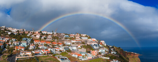 Whitewashed and terracotta homes terrace down steep cliffs toward the Atlantic in Madeira, Portugal, as a vivid rainbow arcs under sunlit clouds in a wide daytime view. © Aerial Film Studio