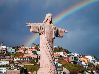 Cristo Rei statue stands on a hillside at Garajau, Madeira, Portugal, arms wide as a rainbow arcs behind. Dramatic cliffs and terraced homes descend toward the sea. © Aerial Film Studio