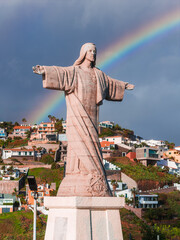 Cristo Rei statue at Garajau, Madeira, Portugal stands on a pedestal as a rainbow arcs after rain,...