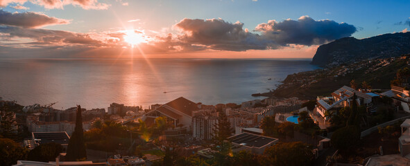 Funchal, Madeira, at sunset, with terraced houses and hotel blocks on steep hills, harbor silhouettes, golden rays on the Atlantic, and a dark headland framing the right © Aerial Film Studio