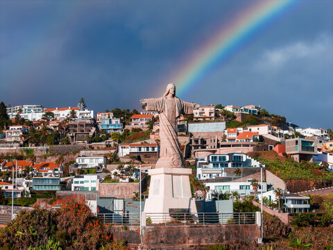 A large Christ the King statue stands above terraced homes in Canico, Madeira, Portugal, as a vivid rainbow arcs behind it and rain cleared light defines the scene.