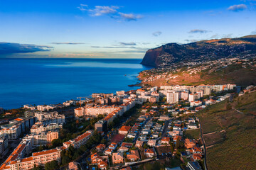 Aerial view of Funchal, Madeira, Portugal shows whitewashed blocks and hotels by the Atlantic, Cabo Girao in distance, warm late afternoon light, clear blue sky. © Aerial Film Studio