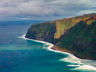 Emerald cliffs drop to the Atlantic near Ponta do Pargo, Madeira. White surf lines rugged rock, a plateau settlement sits above terraces, with moody overcast light.