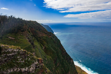 High vantage shows rugged cliff and narrow ledges over foaming surf in Madeira, Portugal, near Cabo Girao, under partly cloudy midday light and wide horizon