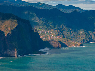 Steep basalt cliffs meet turquoise Atlantic by Madeira villages, late afternoon light reveals terraced hills, winding roads, misty mountains, sea cave near Porto Moniz. © Aerial Film Studio