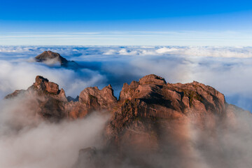 Jagged reddish brown peaks rise above a cloud deck at Pico do Arieiro, Madeira, Portugal. Warm light defines ridgelines as summits appear like islands at sunrise.
