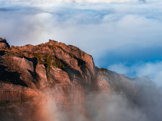 Jagged volcanic ridge rises above an inversion layer near Pico do Arieiro, Madeira, Portugal. Warm low light hits stratified rock as mist drifts, revealing hardy vegetation.