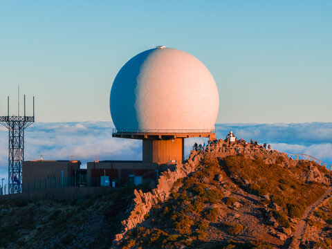 A white radar dome sits on a rugged ridge at Pico do Arieiro, Madeira, Portugal. People stand near a shelter, antenna masts and buildings nearby, warm low light highlights rocks.