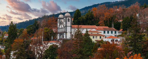 Twin towers rise above terracotta roofs and autumn trees in Monte, Madeira at sunset. Warm light hits whitewashed walls, forested slopes, and soft pink clouds. © Aerial Film Studio