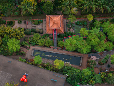 Aerial view shows a red roof pavilion and straight footbridge over a long pond at Monte Palace Tropical Garden in Funchal, Madeira, Portugal in soft early light.