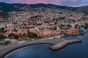 Aerial view of Funchal, Madeira, in evening light, white roofs leading to Praia do Almirante Reis, curved jetty, and Sao Tiago Fortress by the pebble shore. © Aerial Film Studio