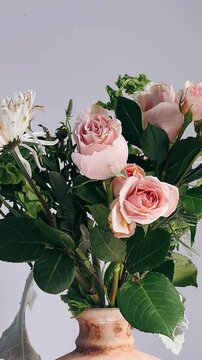 A delicate floral arrangement featuring blush pink roses and a white chrysanthemum surrounded by layered green leaves in a softly textured ceramic vase under natural light.