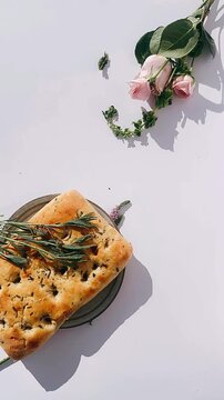 golden rosemary focaccia resting on a plate with scattered herbs and pale pink roses arranged on a clean white background.