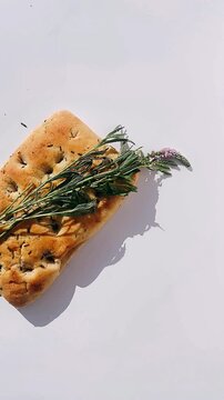 golden focaccia topped with rosemary and herbs resting on a clean white background with gentle shadows