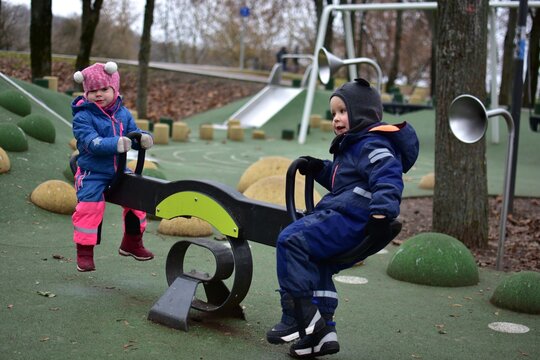 Two toddlers playing together on playground seesaw in autumn park. Children in warm winter outfits enjoy cooperative outdoor play, learning balance and social skills.