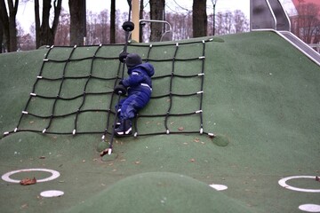 Toddler resting on a playground rope net. Child enjoys playful outdoor activity while developing confidence, balance and motor skills in an autumn park.
