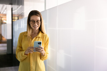 Positive stylish young professional woman in glasses typing on smartphone in office corridor, browsing social media, Internet on work break, enjoying online communication, wireless technology