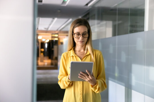 Serious young businesswoman in glasses typing on tablet pc in office corridor, working on Internet project, using digital technology for online business process management, browsing social media - Powered by Adobe