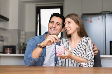 Attractive young couple placing coins into cute piggy bank, start their life together, savings money for purchases, seated at table in modern kitchen. Shared future, responsible planning, partnership