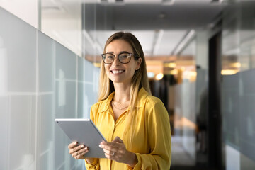 Happy beautiful business professional woman in stylish glasses posing in office corridor with glass walls, holding digital tablet pc, using technology for work, looking at camera, posing for portrait