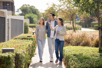 Young professional real estate agent leads smiling couple along landscaped pathway in modern residential complex, speaks about amenities and benefits. Selling contemporary architecture, homeownership
