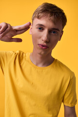 Young man in a yellow shirt makes a peace sign near his head against a bright yellow backdrop,...
