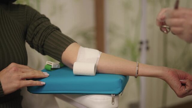 Skilled nurse in medical clinic applying bandage with scissors to patient arm after drawing blood for test, finishing vein puncture procedure with care, close-up. Shooting in slow motion.