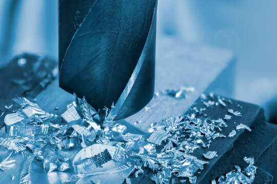 Closeup of steel solid core drill bit working on workpiece in metallic blue tone. Detail of sharp twist cutting tool used to enlarge hole in aluminum profile with metal turnings and blurry background.