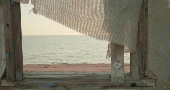 Scenic view of the calm ocean and sandy beach through the weathered wooden window frame of an old, abandoned, and destroyed house, symbolizing hope, memory, and new beginnings.