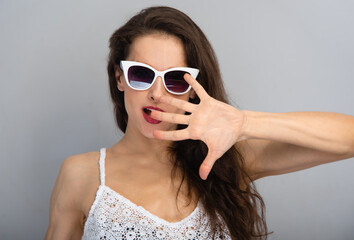 Fototapeta premium Glamour red lipstick smiling fashion woman posing in fashion sunglasses and covers her face with her palm on white wall background with copy space. Lifestyle casual portrait