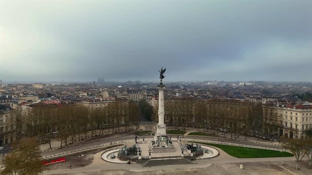 Drone shot of Monument aux Girondins on Esplanade des Quinconces under overcast sky, with angel statue above trees and historic buildings