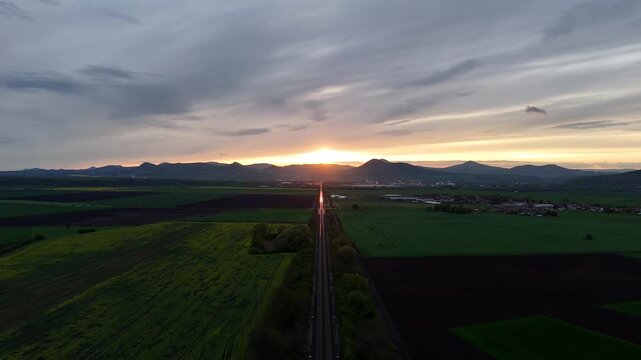 Drone aerial view of railway leading toward mountains at sunset, with a train moving into the distance and sunlight reflecting on rails in a cinematic landscape scene.