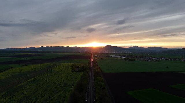 Drone aerial view of railway leading toward mountains at sunset, with a train moving into the distance and sunlight reflecting on rails in a cinematic landscape scene.