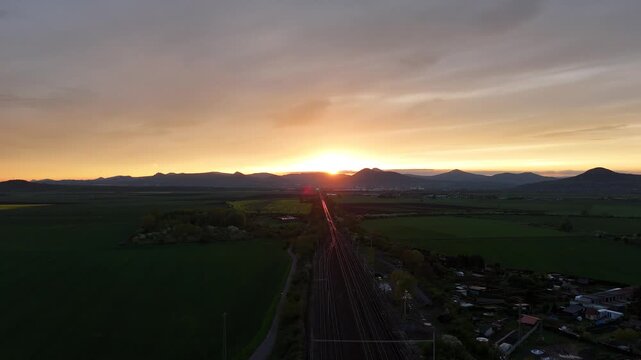 Drone aerial view of railway leading toward mountains at sunset, with a train moving into the distance and sunlight reflecting on rails in a cinematic landscape scene.