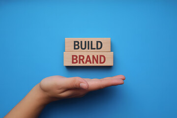 Minimalist flat lay of wooden blocks with BUILD and BRAND on bright blue background, featuring a hand presenting the blocks. Soft lighting enhances warm wood texture.